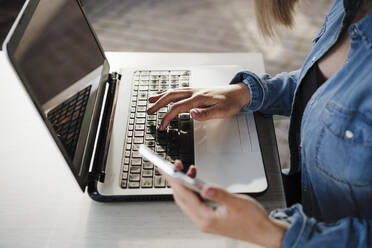 Female entrepreneur holding mobile phone while working on laptop at coffee shop - EBBF03376