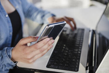 Female professional holding mobile phone while working on laptop in cafe - EBBF03378