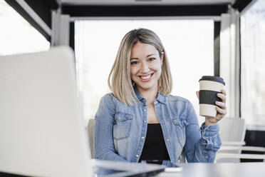 Happy businesswoman holding disposable coffee cup while sitting with laptop at cafe - EBBF03385