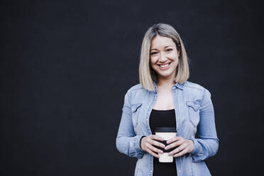 Geschäftsfrau lächelt, während sie eine Einweg-Kaffeetasse vor einer schwarzen Wand hält - EBBF03388