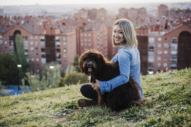 Smiling blond woman embracing dog while sitting on grass - EBBF03391