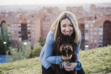 Smiling woman with blond hair embracing Spanish Water Dog on grass during sunset - EBBF03392