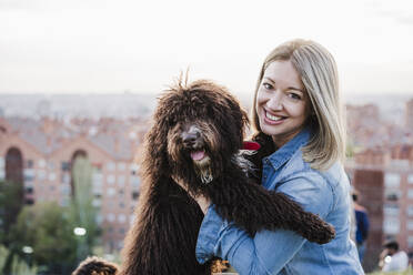 Lächelnde erwachsene Frau mit Hund vor einer Stadtlandschaft - EBBF03393