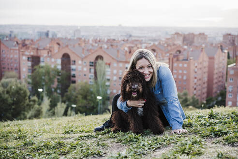 Glückliche Frau, die einen spanischen Wasserhund vor einer Stadtlandschaft umarmt - EBBF03394