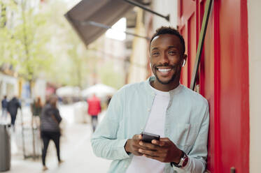 Smiling man with smart phone leaning on red wall - PGF00529