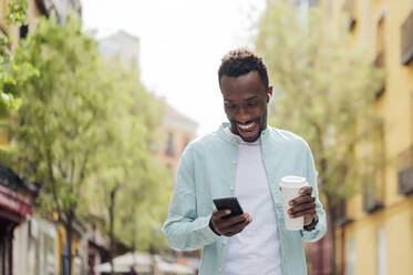 Smiling young man with reusable cup using smart phone at street - PGF00532