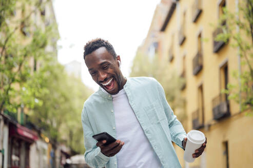 Laughing young man with reusable cup using smart phone at street - PGF00533