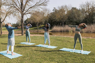 Male yoga instructor guiding to young women while doing stretching exercise on mat during sunny day - JRVF00459