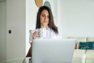 Mittlere erwachsene Frau hält eine Kaffeetasse, während sie zu Hause einen Laptop benutzt - KIJF03782