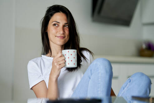 Nachdenklich lächelnde Frau, die mit einer Kaffeetasse zu Hause sitzt - KIJF03787