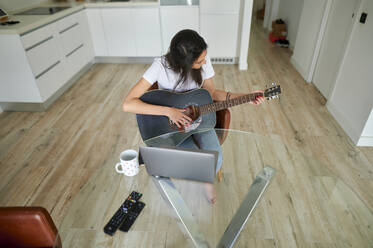 Mittlere erwachsene Frau übt Gitarre, während sie mit einem Laptop zu Hause sitzt - KIJF03791