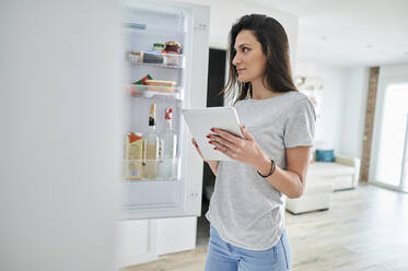 Mittlere erwachsene Frau prüft Lebensmittel im Kühlschrank mit Tablet zu Hause - KIJF03796