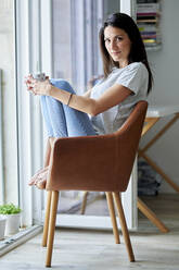 Beautiful woman holding coffee cup while sitting on chair at home - KIJF03804