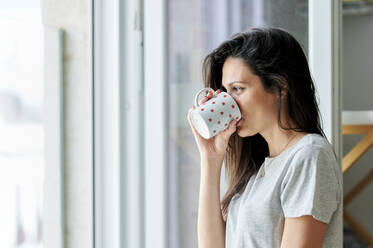 Woman looking through window while drinking coffee at home - KIJF03807