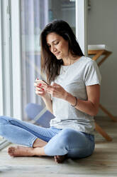 Lächelnde Frau, die ein Mobiltelefon benutzt, während sie zu Hause am Fenster sitzt - KIJF03808