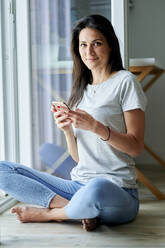 Smiling woman using mobile phone while sitting by window at home - KIJF03809