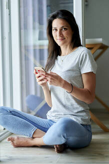 Lächelnde Frau, die ein Mobiltelefon benutzt, während sie zu Hause am Fenster sitzt - KIJF03809