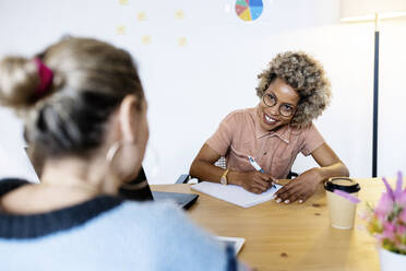 Businesswoman discussing with female colleague while writing in book at home office - RFTF00056