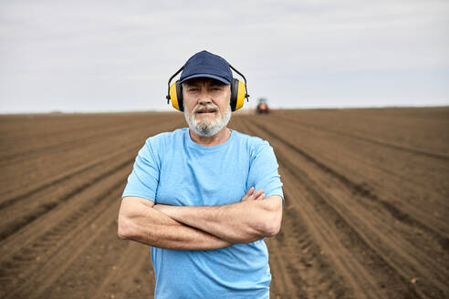 Männlicher Landwirt mit Gehörschutz, der mit verschränkten Armen auf einem landwirtschaftlichen Feld steht - ZEDF04034