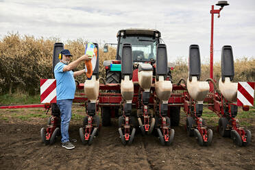 Männlicher Landwirt mit Ohrenschützern beim Einfüllen von Mais in die Behälter eines Traktors auf einem landwirtschaftlichen Feld - ZEDF04040