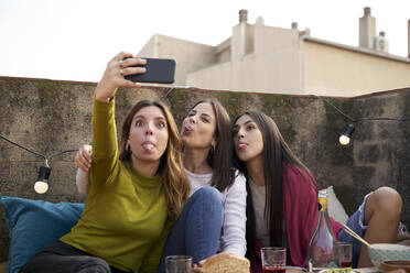 Female friends sticking out tongue while taking selfie on rooftop - VEGF04258