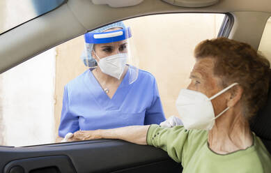 Female doctor wearing protective workwear while looking at senior woman in car - JCCMF01982