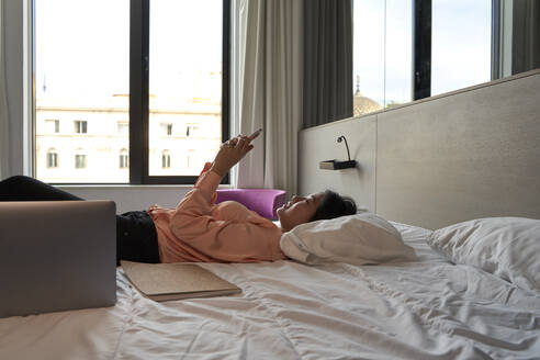 Weibliche Berufstätige, die mit ihrem Smartphone Textnachrichten verschickt, während sie auf dem Bett im Hotel liegt - VEGF04319
