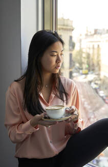 Geschäftsfrau hält Kaffeetasse und blickt durch das Fenster eines Hotels - VEGF04323