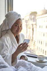 Junge Frau im Bademantel hält Handy am Fenster eines Hotels - VEGF04328