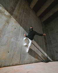 Unbekannter junger Mann in modischem Outfit und Maske, der in einem verlassenen Gebäude an einem sonnigen Tag an einer Betonwand tanzt - ADSF23104