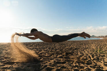 Ganzkörper-Seitenansicht eines hemdlosen, nicht erkennbaren Sportlers, der einen Sprung ausführt und in der Luft über dem Strand mit fliegendem Sand schwebt, während eines Trainings im Freien - ADSF23125