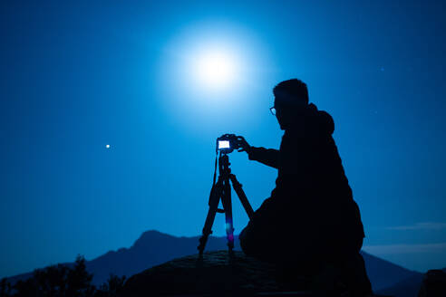 Seitenansicht einer nicht erkennbaren männlichen Reisenden-Silhouette mit Fotokamera auf einem Stativ gegen einen Bergrücken unter blauem Himmel mit glänzender Sonne bei Nacht - ADSF23130