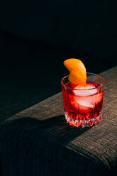 Ein Glas erfrischender Negroni-Cocktail mit bitterem Geschmack und Eis, garniert mit Orangenschale und serviert auf dem Arm der Couch in einem dunklen Raum - ADSF23135