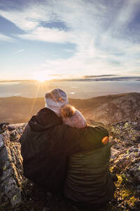 Rückenansicht eines nicht erkennbaren Mannes, der seine Freundin in Oberbekleidung umarmt, während er die Morgendämmerung auf einem Berg bei bewölktem Himmel und Sonnenschein bewundert - ADSF23144