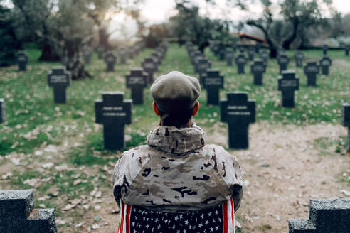 Rückenansicht eines Soldaten in Uniform, der auf einem Stuhl mit amerikanischer Flagge sitzt, während er den Tod von Kriegern auf einem Friedhof betrauert - ADSF23149