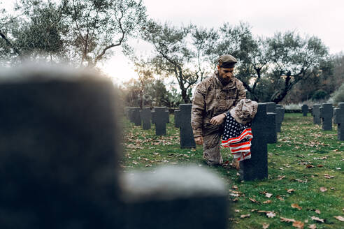 Ganzkörper trauernder Soldat in Tarnkleidung kniet vor einem Grab auf einem Militärfriedhof am frühen Herbsttag - ADSF23150