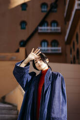 Junge Frau, die ihre Augen abschirmt, während sie an einem sonnigen Tag vor einem Gebäude steht - TCEF01794