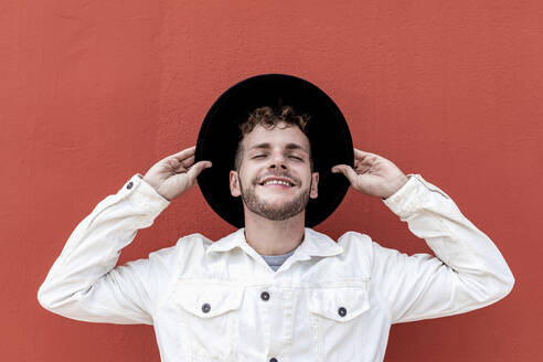 Fröhlicher männlicher Millennial in modischer Kleidung, der seinen Hut zurechtrückt und mit geschlossenen Augen lächelt, während er sich auf der Straße an einer roten Wand ausruht - ADSF23156