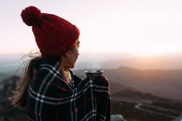 Seitenansicht eines Wanderers in einer Decke, der heißen Kaffee trinkt und auf einem Stein steht und die erstaunliche Landschaft des Hochlandtals an einem sonnigen Tag beobachtet - ADSF23166