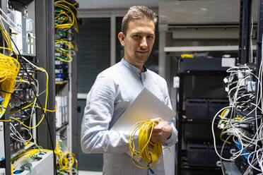 Positiver Mann mit drahtlosem Headset, stehend mit Netbook und Kabeln im Serverraum - ADSF23173