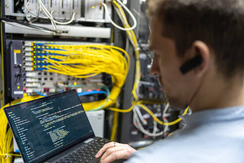 Back view of crop male assistant putting cable in router while using laptop for checking network system - ADSF23174