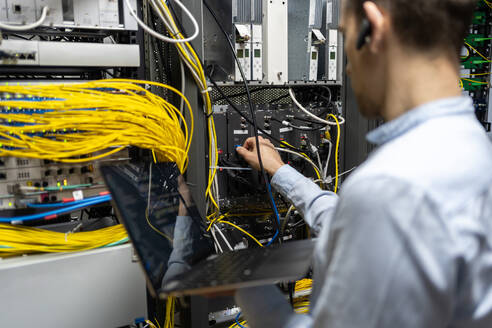 Back view of crop male assistant putting cable in router while using laptop for checking network system - ADSF23175