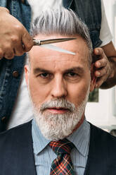 Ein gesichtsloser Friseur schneidet das graue Haar eines selbstsicheren, eleganten männlichen Kunden mittleren Alters mit Bart in Anzug und Krawatte - ADSF23176