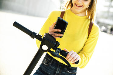 Junge Frau, die ihr Smartphone benutzt, um einen elektrischen Motorroller zu entriegeln - DAF00031