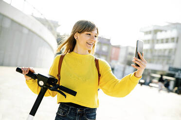 Lächelnde junge Frau nimmt Selfie durch Handy während sonnigen Tag - DAF00034