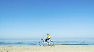 Young woman riding bicycle at beach on sunny day - DAF00038