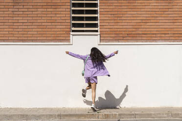 Mid adult woman with arms outstretched dancing in front of wall during sunny day - AFVF08637