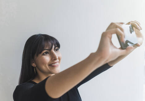 Lächelnde mittelgroße erwachsene Frau, die ein Selfie mit ihrem Mobiltelefon zu Hause macht - DSIF00413