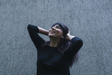 Happy woman with hand in hair looking up while standing in front of wall - DSIF00423