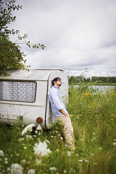 Young man at camper trailer in idyllic lakeside meadow - FSIF05616